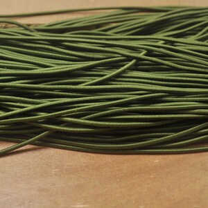 Puede incluir: Primer plano de un montón de cordón elástico verde oliva. El cordón está enrollado firmemente y parece estar hecho de un material suave y flexible. El fondo es una superficie de color marrón claro.