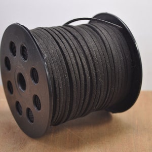 Op de afbeelding: Een spoel met zwart suède koord, gewikkeld om een zwarte plastic spoel met meerdere gaten. Het koord is glad en heeft een licht getextureerd oppervlak.