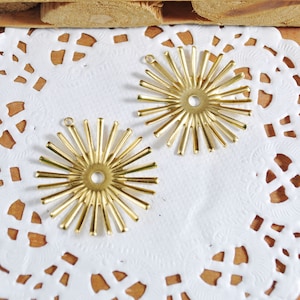 Może przedstawiać: Dwa złote metalowe charms w kształcie rozkwitającego słońca z okrągłym otworem w środku. Charms leżą na białym obrusie.
