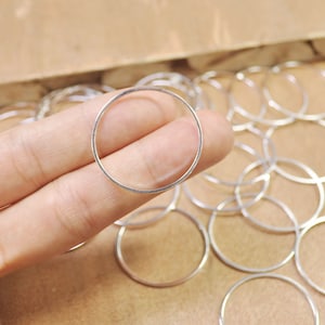 May include: A close-up of a silver jump ring, with many other jump rings in the background. The jump ring is held in a hand.