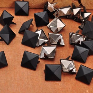 May include: A collection of black square pyramid-shaped studs with silver metal prongs. These decorative studs are scattered on a brown surface. The studs are likely used for embellishing clothing, accessories, or other craft projects.