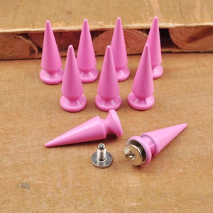 May include: A collection of pink cone-shaped studs, each with a flat base and a pointed top. The studs are arranged on a wooden surface, with some standing and others lying down, alongside a screw and metal backing.