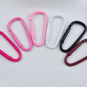 May include: Seven metal keychains in various colors, including pink, white, black, and maroon. The keychains are all in the shape of a simple oval loop.