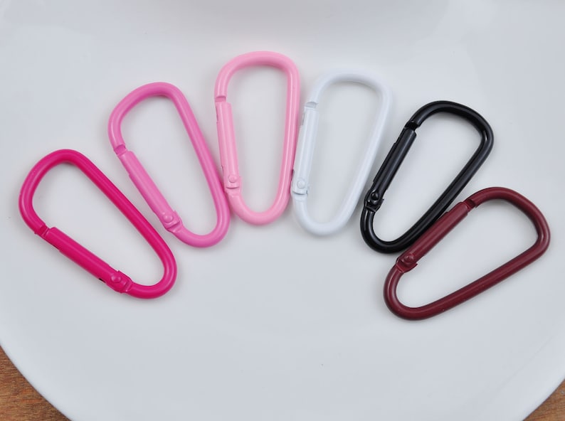 May include: Seven metal keychains in various colors, including pink, white, black, and maroon. The keychains are all in the shape of a simple oval loop.