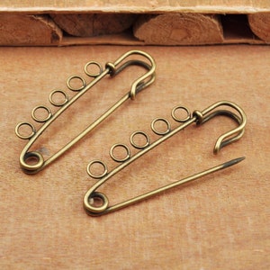 May include: Two antique gold safety pins with multiple decorative loops. The pins are open and lying on a wooden surface.