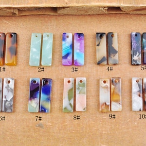 Puede incluir: Una colección de diez pares de colgantes rectangulares en varios colores y patrones. Los colgantes están numerados del 1 al 10. Los colores incluyen marrón, verde, azul, morado y blanco. Cada colgante tiene un agujero en la parte superior para ensartar.