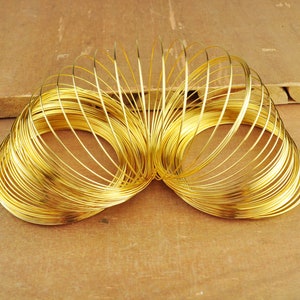 Op de afbeelding: Een gouden metalen spiraalveer, gemaakt van meerdere verbonden ringen, ligt op een houten oppervlak. De veer bevindt zich in een ontspannen, open positie, waarbij de ringen een losse spiraal vormen.