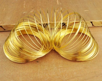 May include: A gold-colored metal coil spring, made of multiple connected rings, is lying on a wooden surface. The spring is in a relaxed, open position, with the rings forming a loose spiral.