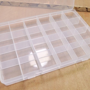 Puede incluir: Una caja de almacenamiento de pl&aacute;stico transparente con 24 compartimentos. La caja est&aacute; abierta y los compartimentos est&aacute;n vac&iacute;os.