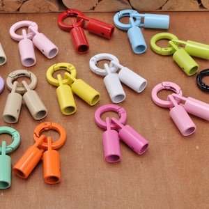 Puede incluir: Una colección de cierres metálicos para cordones en varios colores, cada uno con un anillo circular de resorte unido a dos extremos cilíndricos. El conjunto incluye varios colores sólidos como rojo, azul, amarillo, verde, rosa, blanco, negro y beige.