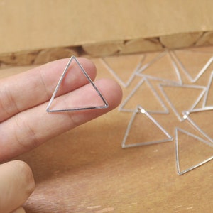 Puede incluir: Un colgante de triángulo de plata. El colgante se sostiene en una mano y hay varios otros colgantes de triángulo esparcidos sobre una superficie de madera.