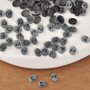 May include: A collection of oval-shaped cameo beads in shades of gray and black, scattered on a white plate. The beads feature a classical profile design. Some beads have a textured surface.