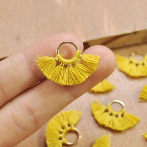May include: A close-up of a yellow tassel with a gold ring. The tassel is made of thin strands of yarn and is shaped like a fan. The ring is attached to the top of the tassel and is used to attach it to a piece of jewelry or other item.