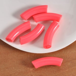 Op de afbeelding: Zes roze gebogen plastic kralen, elk met een gat aan beide uiteinden, op een wit bord.