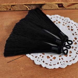 May include: Four black tassel trims displayed on a white lace doily. The tassels are made from soft, silky threads and have a knotted top.
