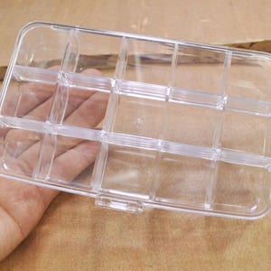 May include: A clear plastic storage box with multiple compartments. The box is empty and has a rectangular shape.