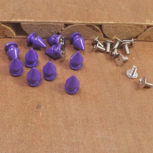 May include: Purple metal spikes with a pointed tip and a flat base. The spikes are arranged in a semi-circle on a wooden surface. There are also several silver screws with a star-shaped head.