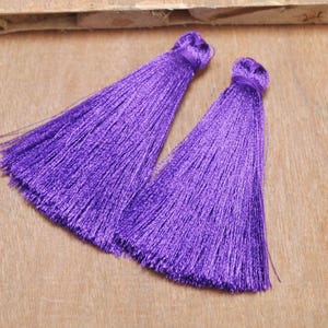 May include: Two purple silk tassels with a long fringe. The tassels are on a wooden surface.