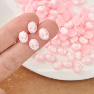 May include: Pink oval cameo cabochons with white relief portraits. Several are held between fingers, with many more scattered on a white surface. These small, flat-backed resin pieces are ideal for jewelry making and crafting.