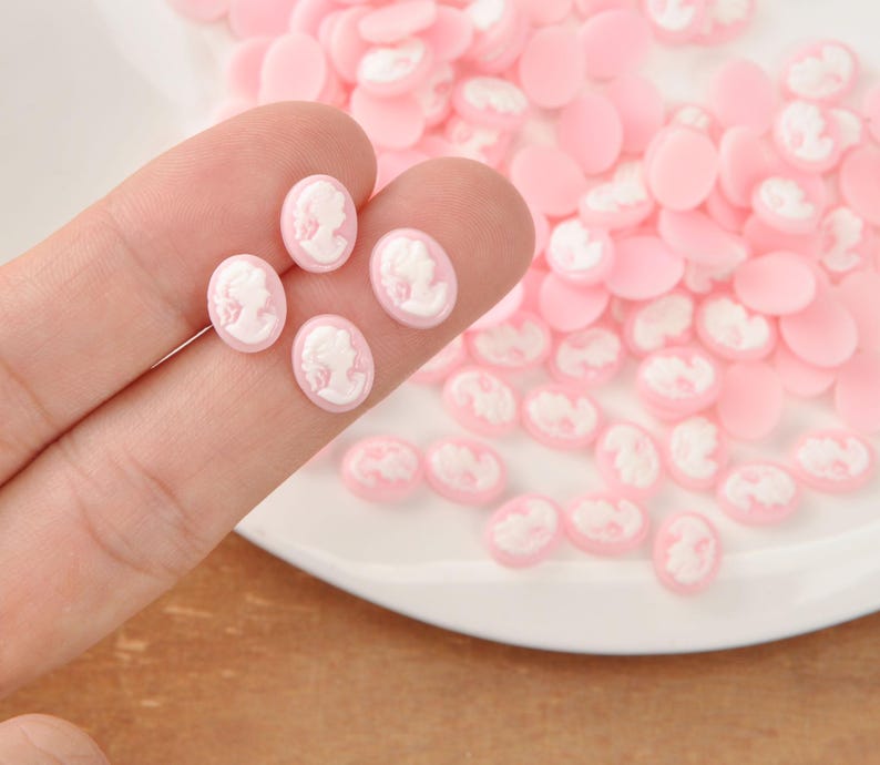 May include: Pink oval cameo cabochons with white relief portraits. Several are held between fingers, with many more scattered on a white surface. These small, flat-backed resin pieces are ideal for jewelry making and crafting.