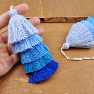 May include: Two ombre tassel trims in shades of blue, light blue, and white. The tassels are made of soft yarn and have a layered, graduated look.