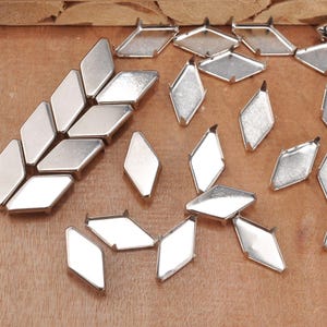 Può includere: Una collezione di componenti per gioielli in metallo argentato a forma di diamante. I componenti hanno una superficie piatta e riflettente e piccole linguette sui lati. Sono sparsi su una superficie di legno.