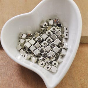May include: A white ceramic heart-shaped dish filled with silver metal cube beads.