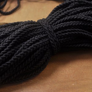Peut inclure: Gros cordon tressé noir, montrant le motif tissé complexe. Le cordon est épais et texturé, avec une sensation douce et souple.
