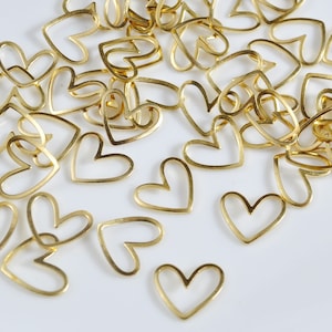 May include: A scattering of gold-colored metal heart-shaped charms. The charms are all the same size and shape, and they are arranged in a random pattern on a white surface.
