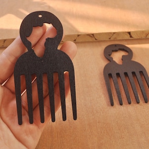Puede incluir: Dos peines afro de madera negra, cada uno con una silueta de persona y peinado afro. Los peines tienen un diseño de peine con varios dientes. Un peine se sostiene en la mano, el otro sobre una superficie de madera. Para peinar el cabello.
