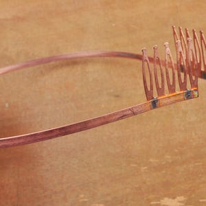 Puede incluir: Una diadema de color cobre con un diseño de peine en el extremo. El peine tiene varios dientes que están espaciados uniformemente.