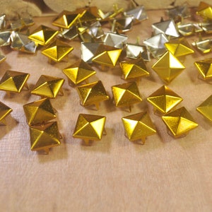 Può includere: Una collezione di borchie metalliche quadrate dorate e argentate con un design a piramide. Le borchie sono sparse su una superficie di legno marrone chiaro. Ogni borchia ha un piccolo perno sul fondo.