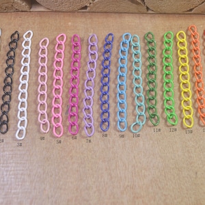 Puede incluir: Quince cadenas metálicas de diferentes colores, cada una de aproximadamente 10 cm de largo, se colocan sobre una superficie de madera. Las cadenas están numeradas del 1 al 15.