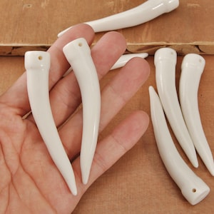 Könnte beinhalten: Gezeigt werden mehrere weiße, gebogene Keramikanhänger, die an Zähne oder Hörner erinnern. Jeder Anhänger hat eine glatte, glänzende Oberfläche und ein kleines Loch oben. Die Anhänger variieren leicht in Größe und Krümmung, was auf eine handwerkliche Qualität hindeutet.
