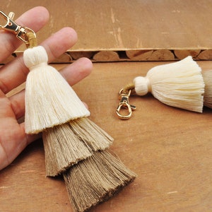 May include: Two-tone tassel keychain with a gold metal clasp. The top tassel is cream coloured and the bottom tassel is brown.