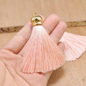 May include: A pair of peach-colored silk tassels with gold metal caps. The tassels are approximately 2 inches long and are held in a hand.