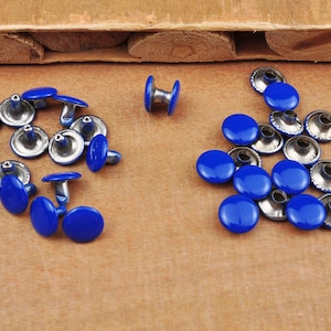 May include: A collection of blue metal rivets with silver accents. The rivets are arranged in two groups on a brown wooden surface.