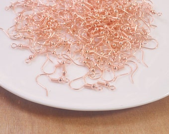 May include: A pile of rose gold earring hooks on a white plate. The hooks are made of metal and have a small loop at the top and a small ball at the bottom.