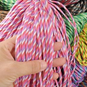 May include: Close-up of colorful paracord bundles. The main focus is on a bundle of pink, red, white, and purple striped paracord. Other bundles in the background include green, yellow, and brown paracord. The paracord is tightly woven and appears durable.