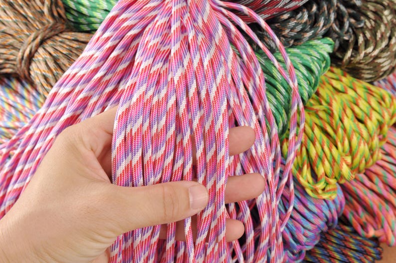 May include: Close-up of colorful paracord bundles. The main focus is on a bundle of pink, red, white, and purple striped paracord. Other bundles in the background include green, yellow, and brown paracord. The paracord is tightly woven and appears durable.