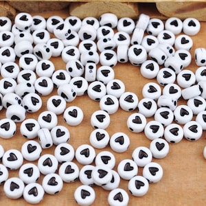 Könnte beinhalten: Eine große Menge weißer runder Perlen mit schwarzen Herzmotiven. Die Perlen sind auf einer Holzoberfläche verstreut.