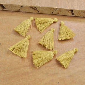 May include: Eight small, mustard yellow tassels with a fringe on a wooden surface.