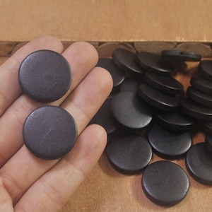 May include: A close-up of a hand holding two black wooden round buttons. A pile of similar buttons is in the background.