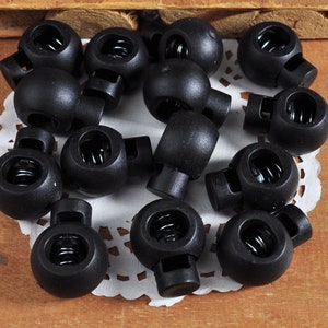 May include: A close-up of a group of black plastic toggle clasps with a spring mechanism. The clasps are arranged on a white lace doily.