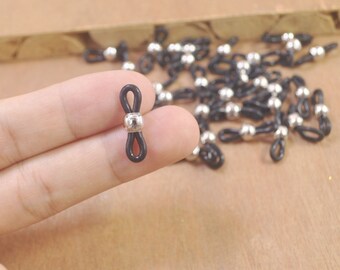 Puede incluir: Primer plano de un cordón de goma negro con una cuenta de metal plateado en el centro. El cordón está anudado en forma de ocho. El cordón es un tipo de soporte para gafas.