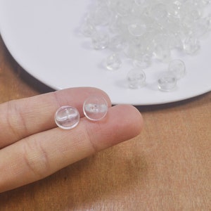 Könnte beinhalten: Zwei durchsichtige Plastikknöpfe, einer in der Hand gehalten, der andere auf einem weißen Teller mit einem Stapel ähnlicher Knöpfe.