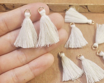 May include: A collection of ten off-white tassels with gold jump rings. The tassels are made of soft, fluffy yarn and are approximately 1 inch in length.