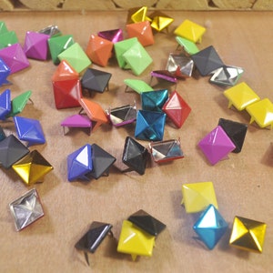 Puede incluir: Surtido de tachuelas metálicas de forma piramidal de colores con acabado brillante. Las tachuelas son de varios colores, incluyendo rojo, verde, azul, amarillo y negro.