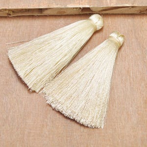 May include: Two ivory silk tassels with a long, thin fringe. The tassels are on a wooden surface.