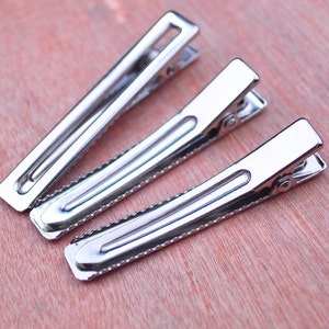 Op de afbeelding: Drie zilverkleurige metalen haarclips diagonaal gerangschikt op een lichtbruin houten oppervlak. De clips hebben een rechthoekige vorm met een scharnierend ontwerp en een gekartelde rand voor grip. De clips zijn van verschillende formaten.